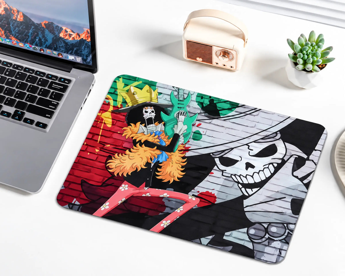 Brook from One Piece wearing a golden crown, orange feathered coat, and pink floral trousers while holding a turquoise guitar, against a red and green graffiti brick wall with a large monochrome skull portrait on a gaming mouse pad.