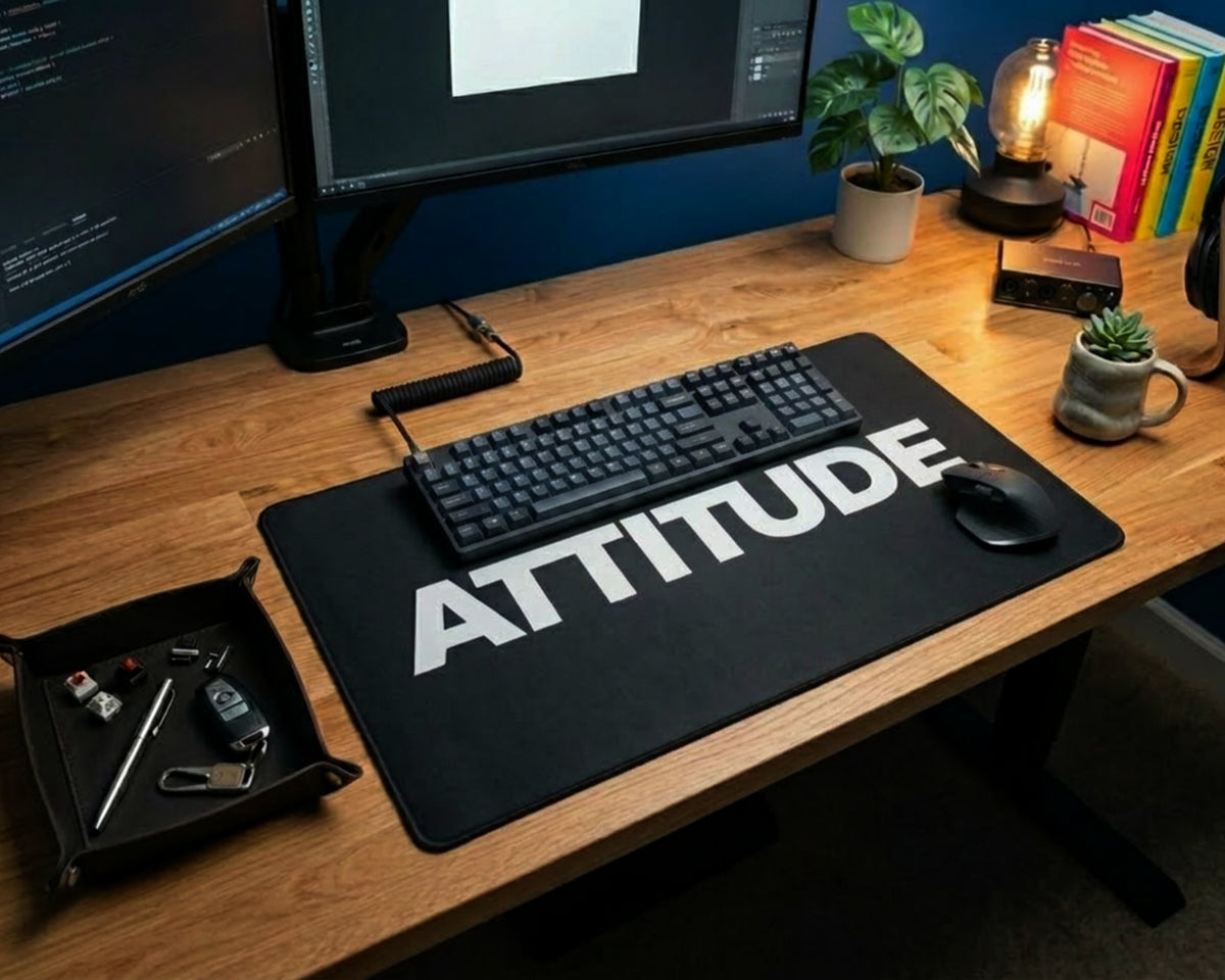Modern wooden desk setup with black attitude desk mat, mechanical keyboard, mouse, dual monitors, plants, books, and warm desk lamp.