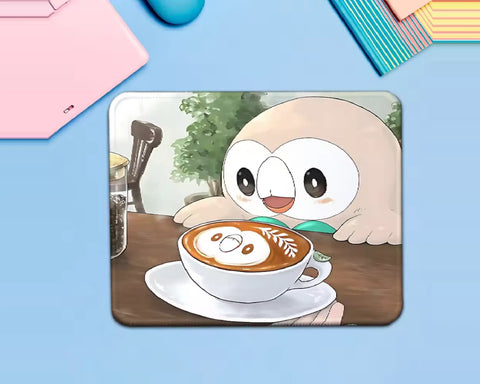 Rowlet Pokémon mouse pad featuring the adorable owl Pokémon looking at a cup of coffee with latte art, set on a wooden café table with a cozy background.